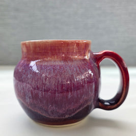 handmade ceramic mug with a handle, featuring a layered glaze transitioning from soft peach tones at the rim into deep plum and rich purple hues toward the base