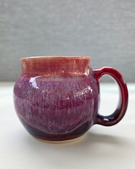 handmade ceramic mug with a handle, featuring a layered glaze transitioning from soft peach tones at the rim into deep plum and rich purple hues toward the base