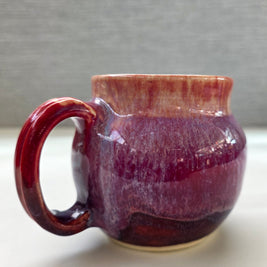 handmade ceramic mug with a handle, featuring a layered glaze transitioning from soft peach tones at the rim into deep plum and rich purple hues toward the base