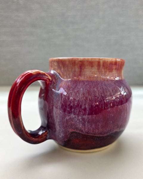 handmade ceramic mug with a handle, featuring a layered glaze transitioning from soft peach tones at the rim into deep plum and rich purple hues toward the base
