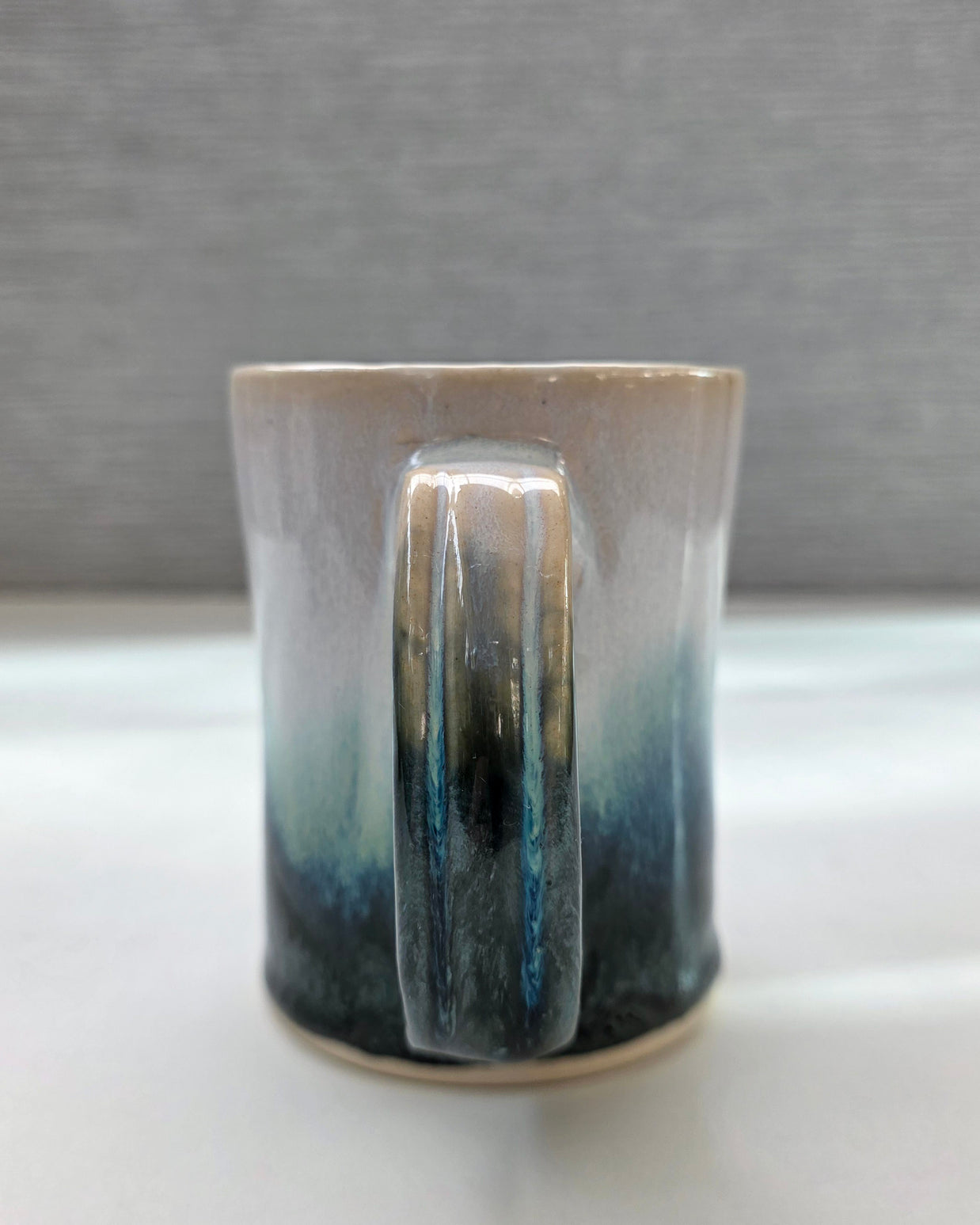 handmade ceramic mug with a handle, finished in a soft gradient glaze transitioning from warm white to a deep teal base
