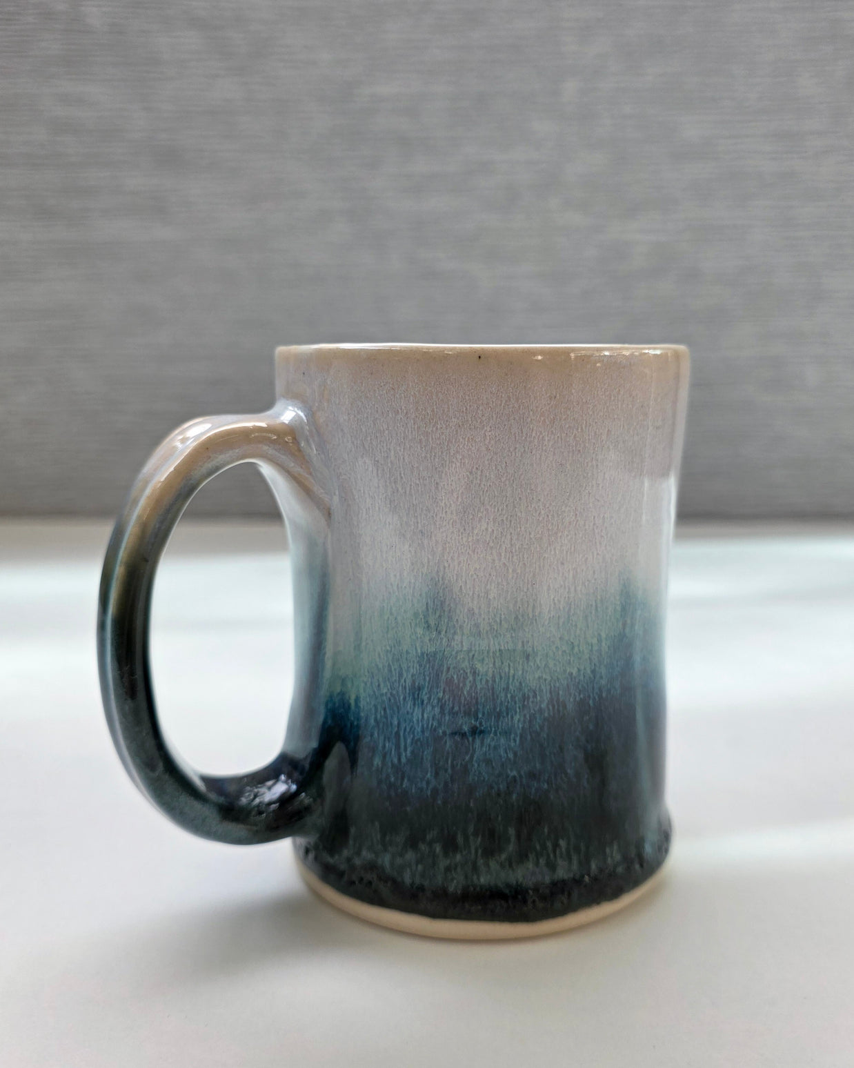 handmade ceramic mug with a handle, finished in a soft gradient glaze transitioning from warm white to a deep teal base